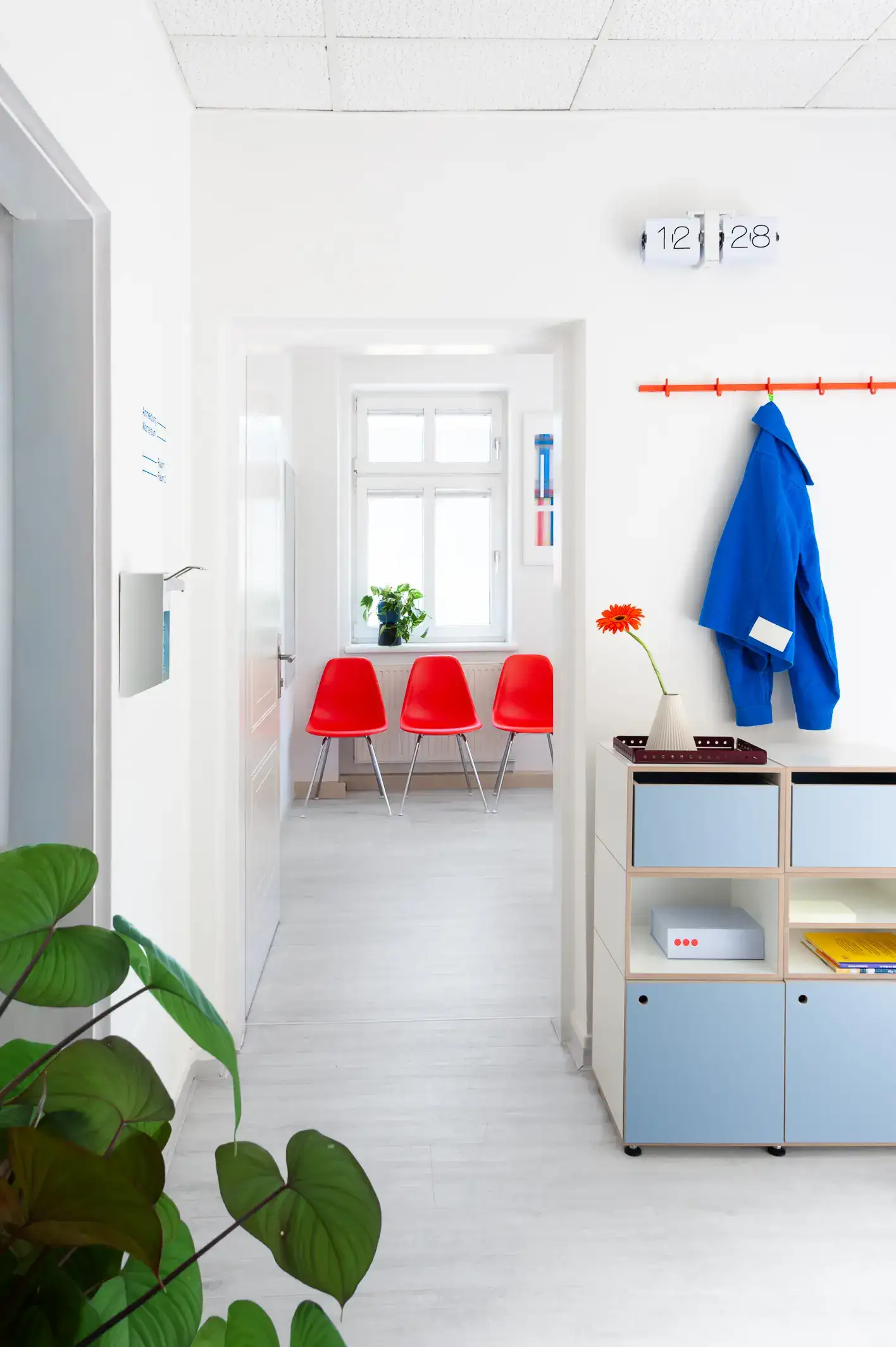 Büro Jutro: Empfangsraum und Wartezimmer in einer Ordination für Allgemeinmedizin View from a reception area with a sideboard in blue into a waiting room with three red chairs and a plant on a window shelf.