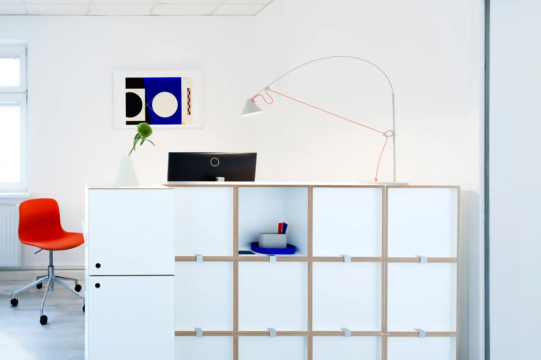 büro jutro: Empfangspult und Raumteiler für eine Ordination für Allgemeinmedizin Reception desk and room divider in a bright general practice made of white modules with a red chair, a lamp, a white vase with a blue flower and a geometric print on the wall.
