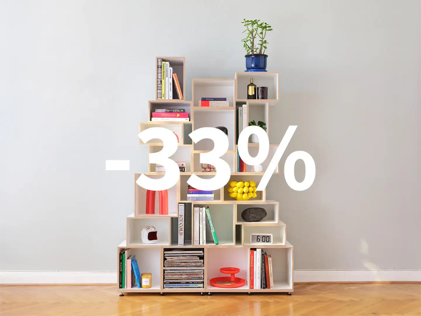 -33 % Rabatt Modern modular living room shelf in a sunlit, airy space. Staggered wooden modules of varying sizes display a curated mix of books, lush green plants, vintage vinyl records, and stylish decor. Set against light grey walls and warm wooden flooring.