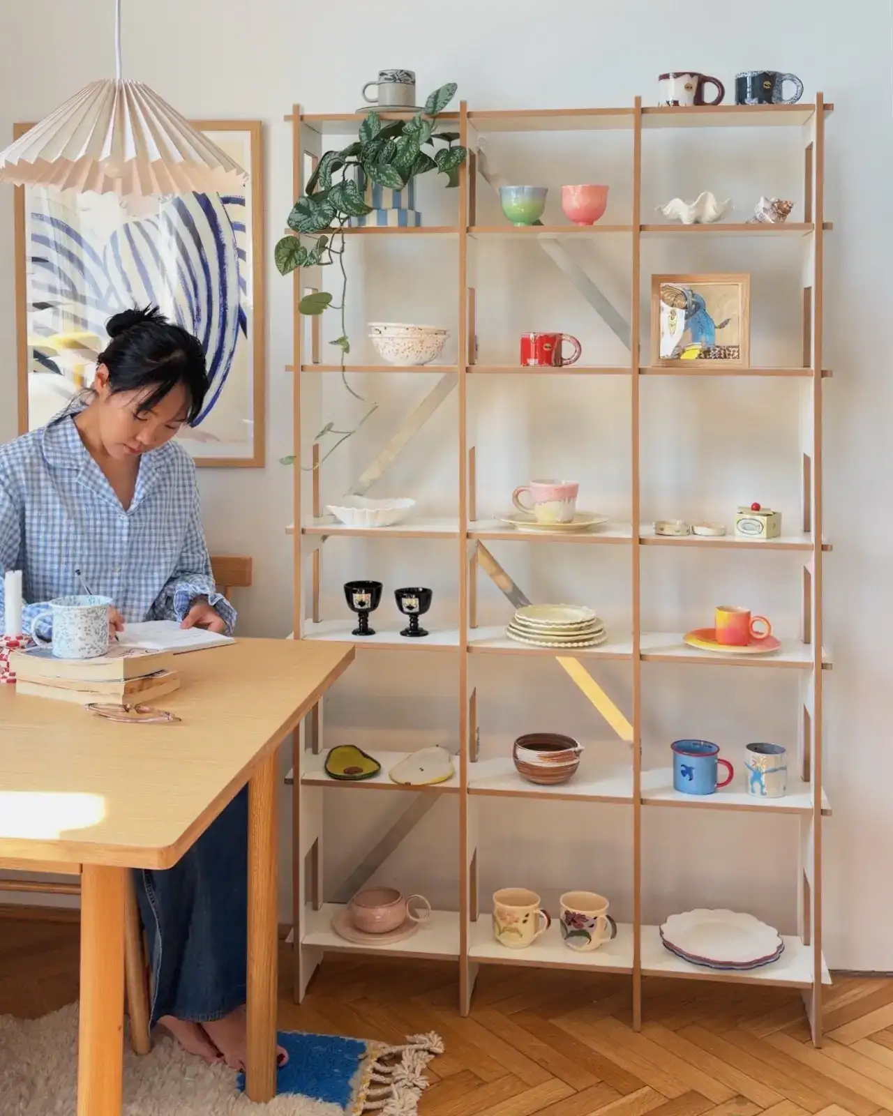 Woman writing in notebook at home desk workspace next to a Nordic-style narrow shelving with white ceramic cups, bowls and a succulent.