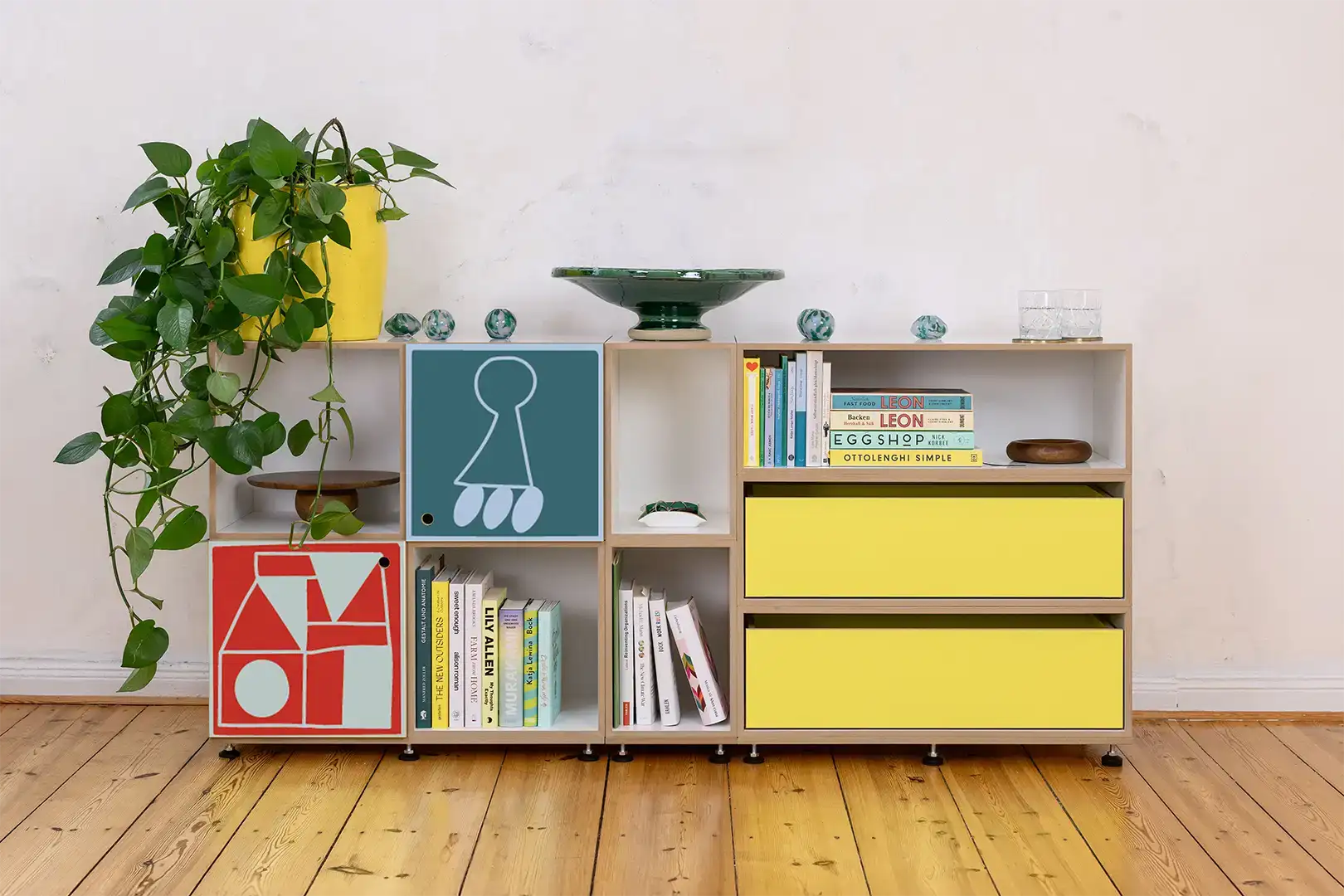 A stylish modular sideboard featuring vibrant yellow drawers and two doors with a bold, colorful graphic motif, set against a light-colored wall and natural wooden flooring  and styled with green plants, books, and small decorative objects
