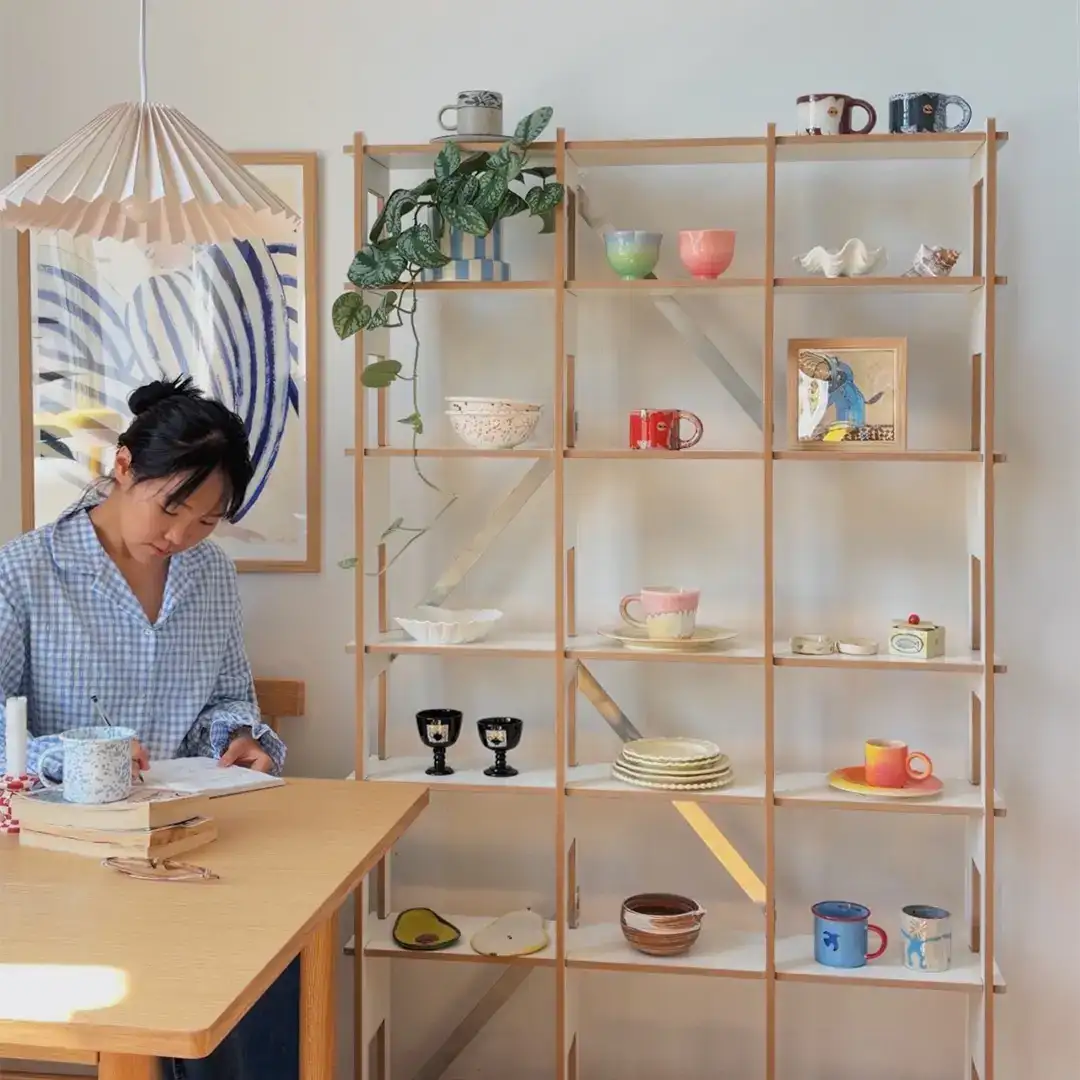 stocubo x mengmengliving Woman writing in notebook at home desk workspace next to a Nordic-style narrow shelving with white ceramic cups, bowls and a succulent.